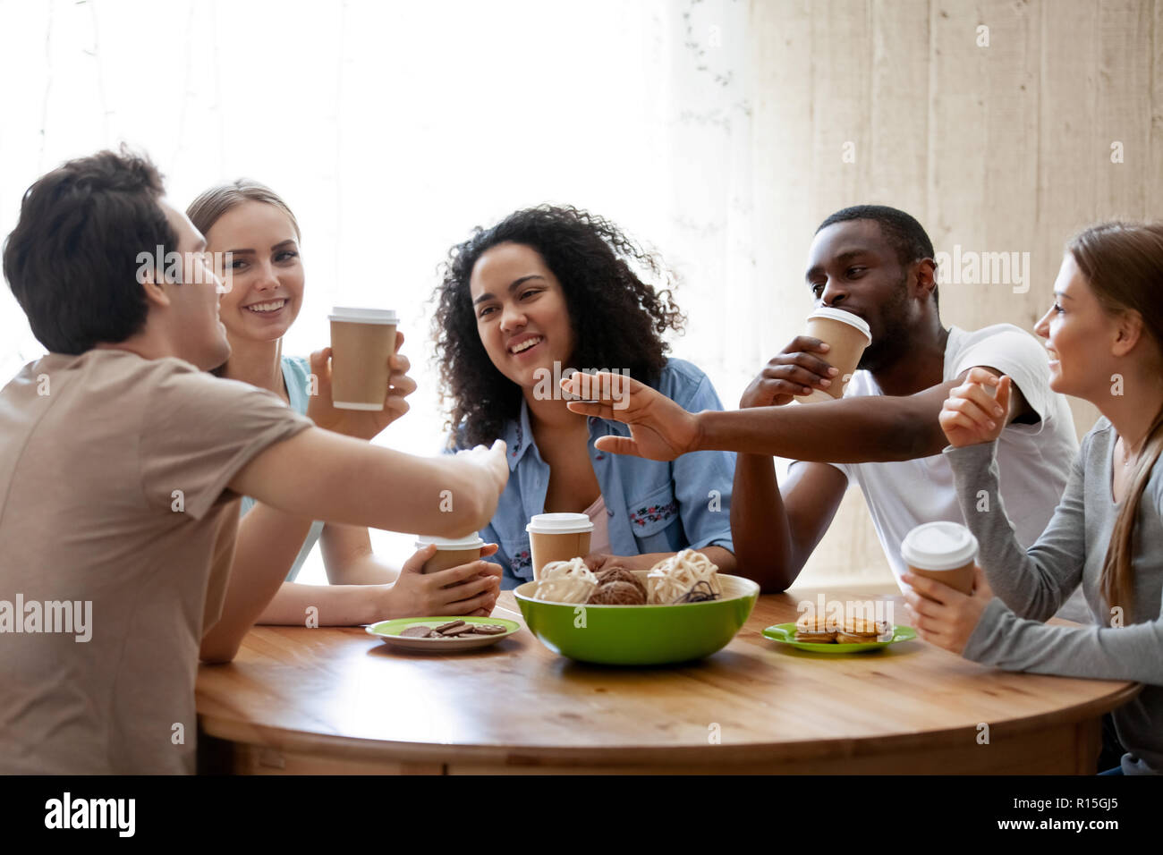 Diverse friends girls and guys sitting around table chatting having fun drink coffee in paper cups enjoy time together. Friendship between different r Stock Photo