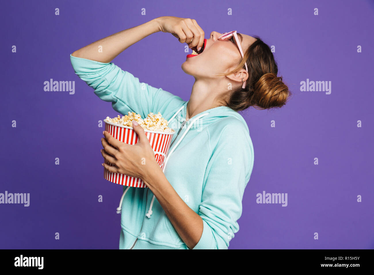 Portrait of a funny young girl with bright makeup isolated over violet background, eating popcorn Stock Photo