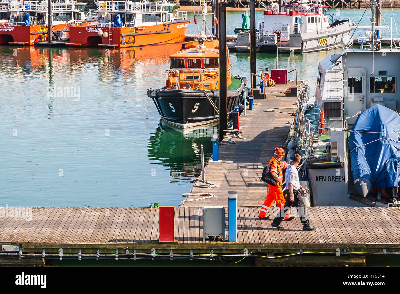 2 workmen walking down the jetty at Ramsgate Harbour with boats and yachts moored in the port Stock Photo