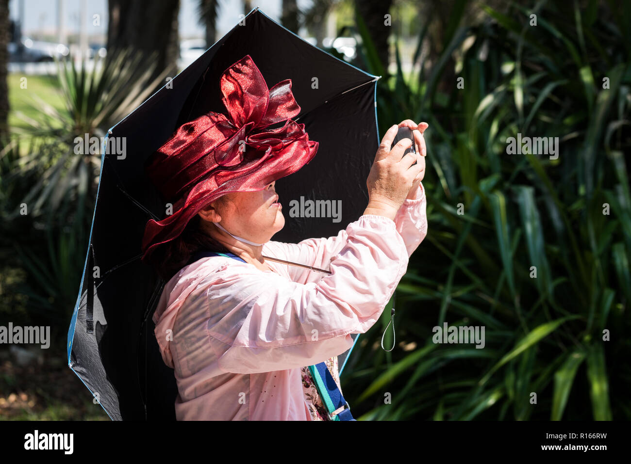 An Asian senior woman wearing bright red hat and holding umbrella while taking picture at Heritage village, Abu Dhabi Stock Photo