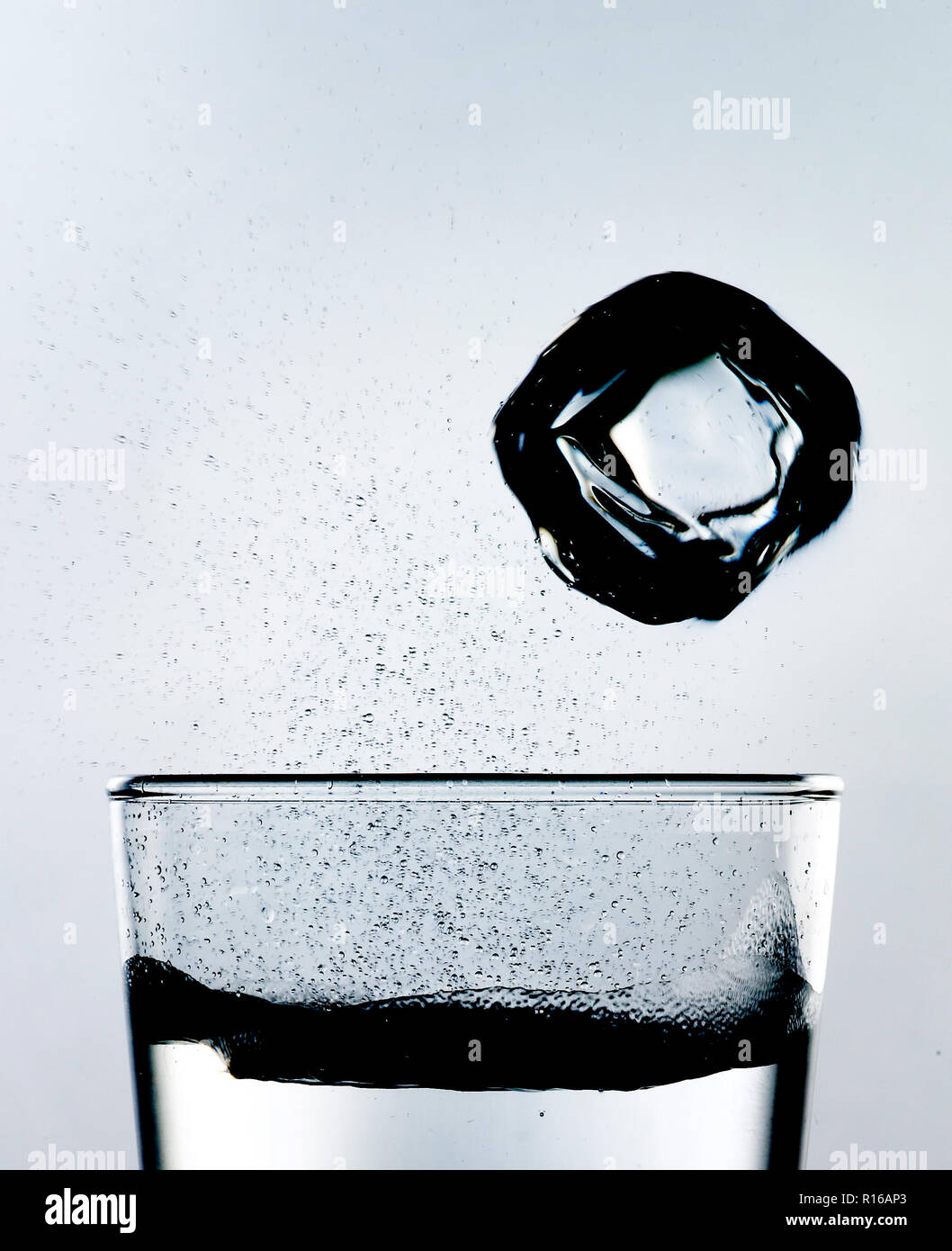 Ice cube falling toward a glass of sparkling effervescent liquid, studio shot, high key Stock Photo