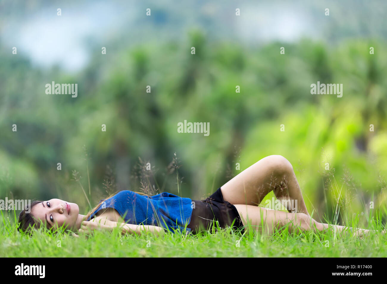 Beautiiful young Filipina woman relaxing on green grass lying on her back in a lush green park looking at the camera with a serene smile Stock Photo