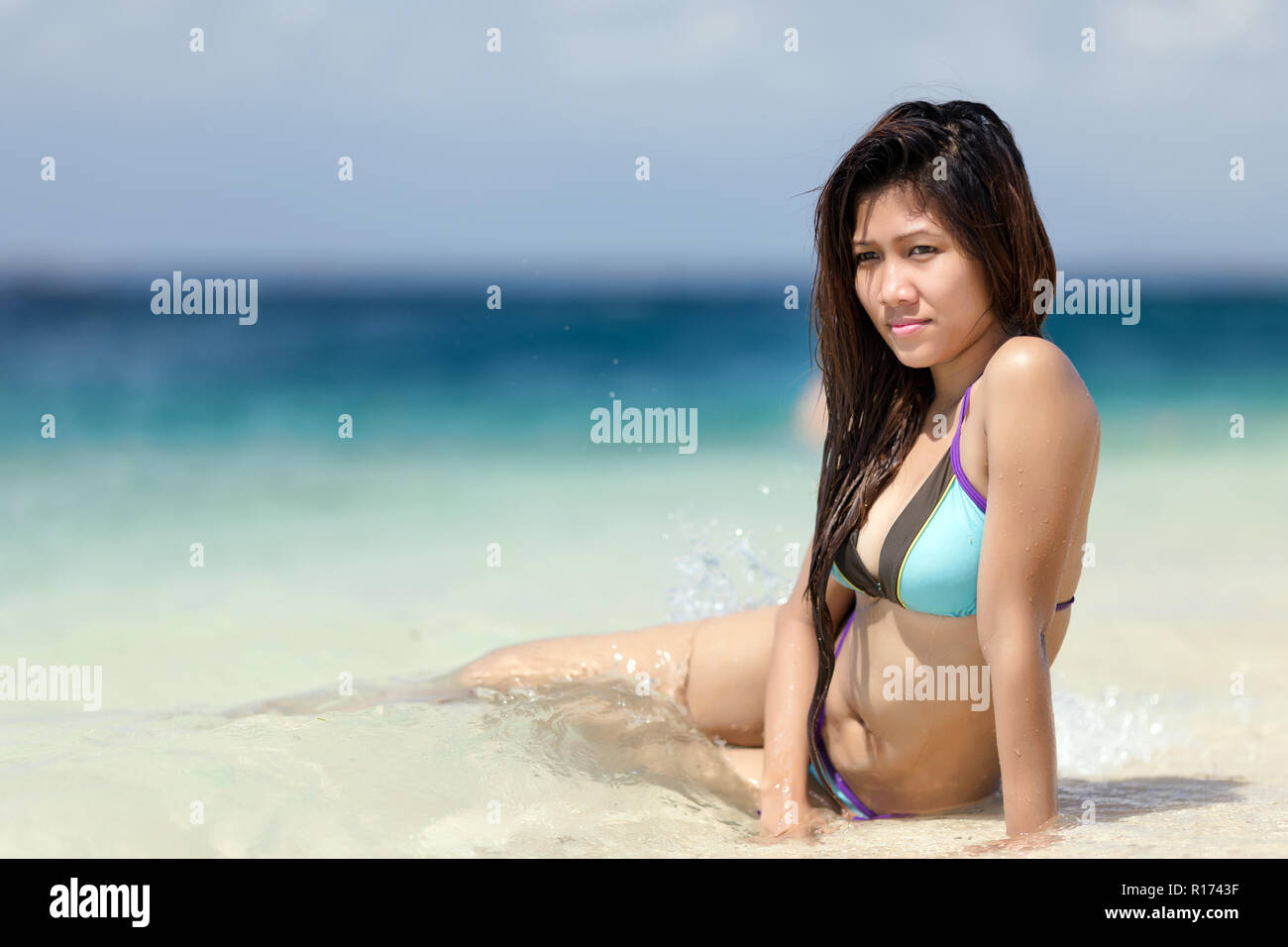 Beautiful young Filipina woman at the seaside sitting in the shallow tranquil blue ocean in a bikini enjoying the hot tropical summer sun, with copysp Stock Photo