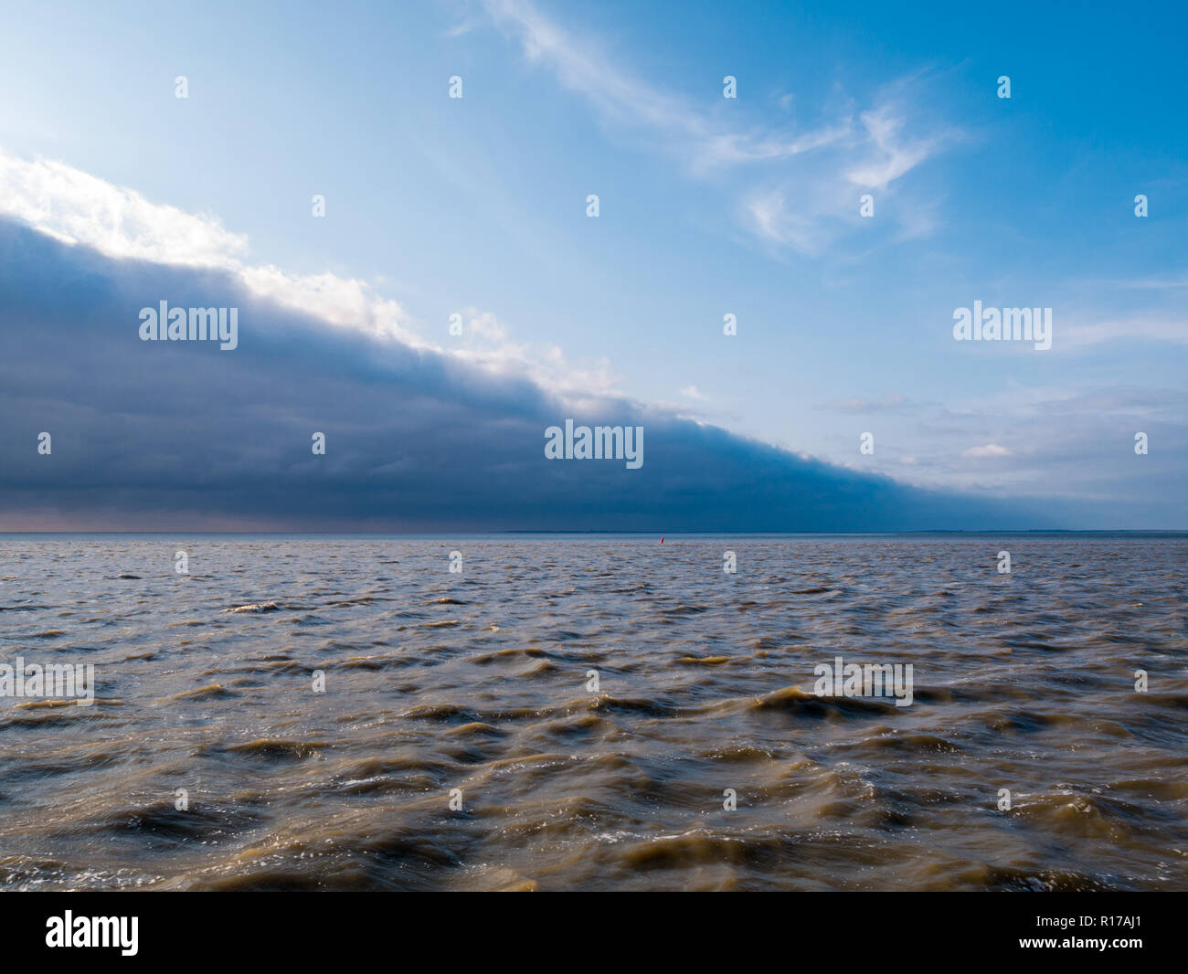 Leading edge with line of storm clouds of cold weather front approaching blue sky on rough water of Wadden sea, Netherlands Stock Photo