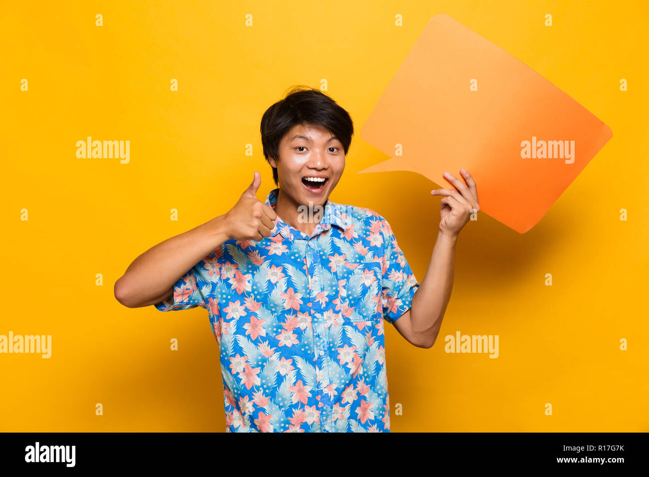 Image of happy young asian man standing isolated over yellow background holding speech bubble showing thumbs up. Stock Photo