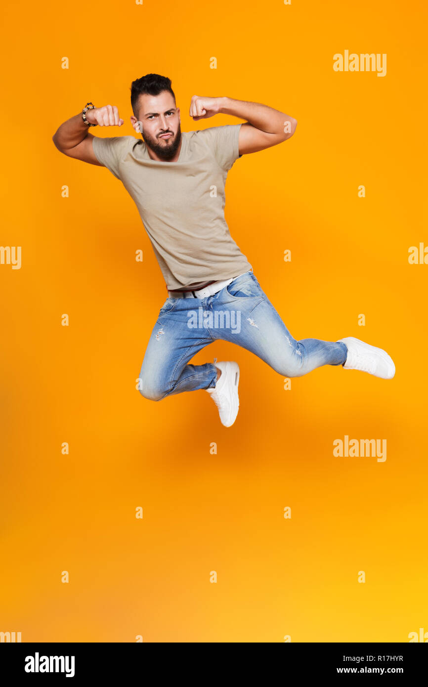 Full length portrait of a smiling young casual man jumping isolated over orange background, flexing biceps Stock Photo