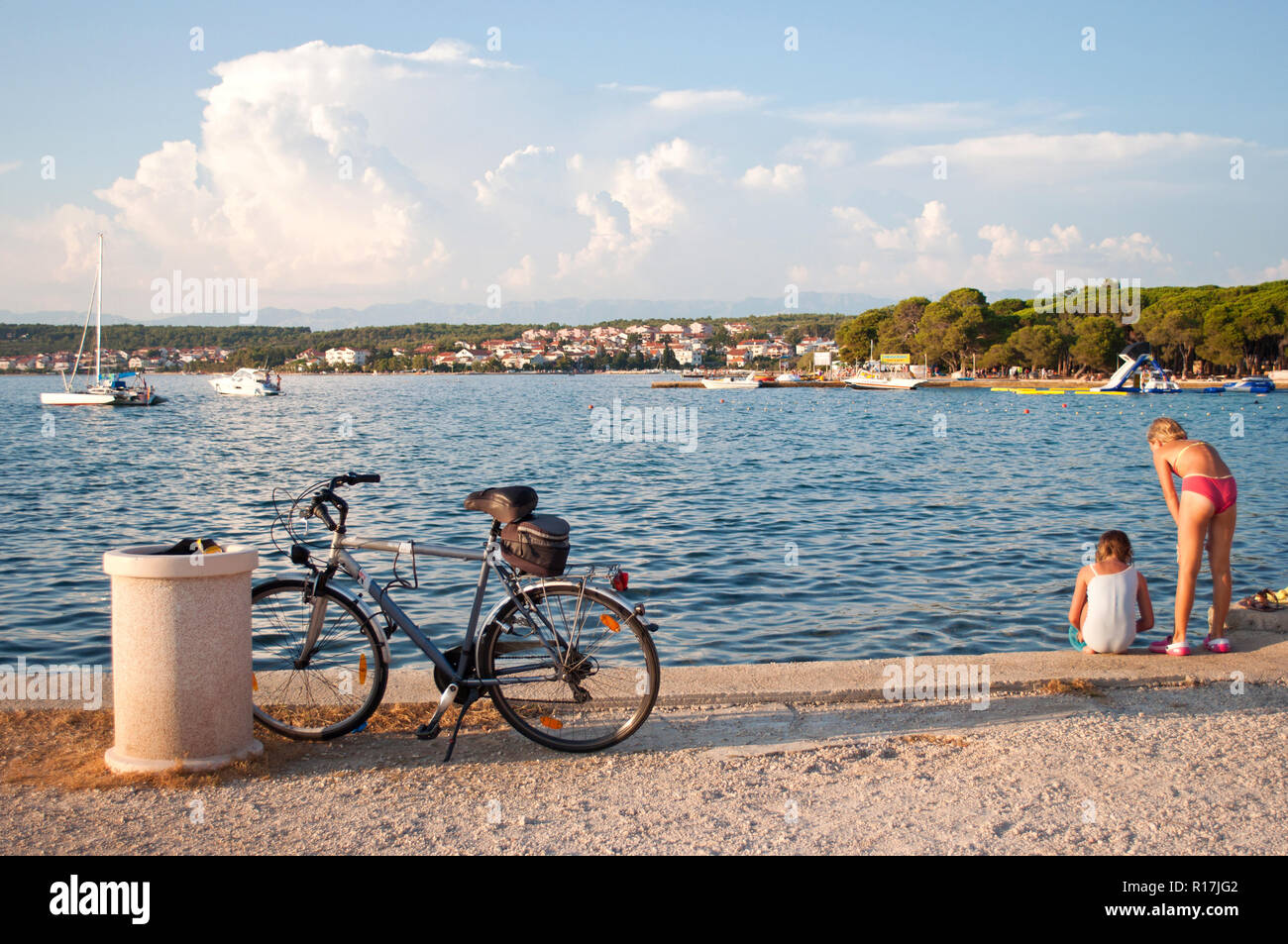 Two girls playing on a sea shore near a bicycle. View on a cozy little Croatian town Zadar covered in white fluffy clouds Stock Photo