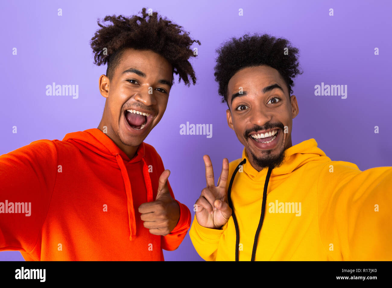 Two happy african man friends dressed in colorful hoodies standing isolated over violet background, taking a selfie Stock Photo