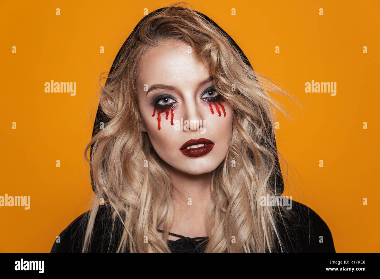 Serious blonde witch wearing black cloak looking camera with scary make up isolated over orange Stock Photo