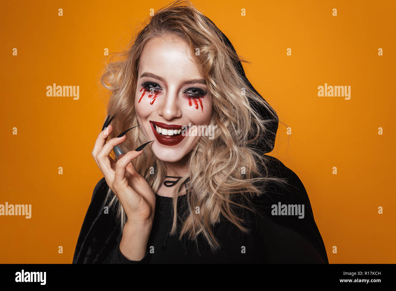 Smiling blonde scary woman witch in black cloak looking camera isolated over orange Stock Photo
