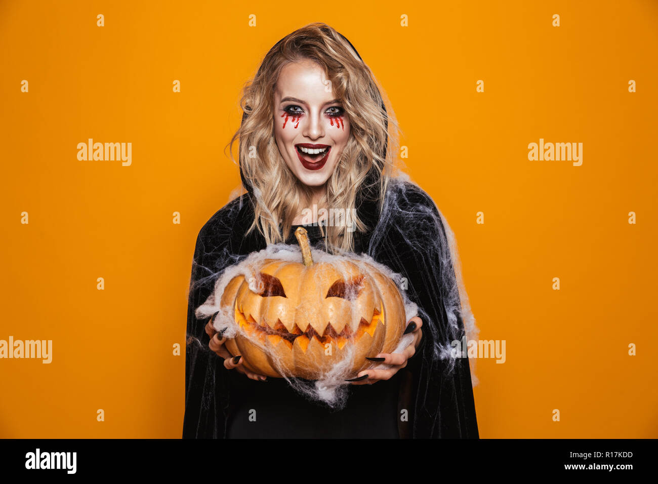 Happy scary blonde witch in black cloak holding pumpkin and laughing to camera isolated over orange Stock Photo