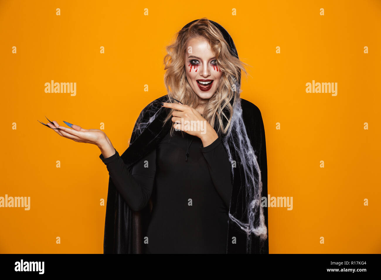 Cheerful scary blonde woman witch pointing at copy space and smiling to camera isolated over orange Stock Photo