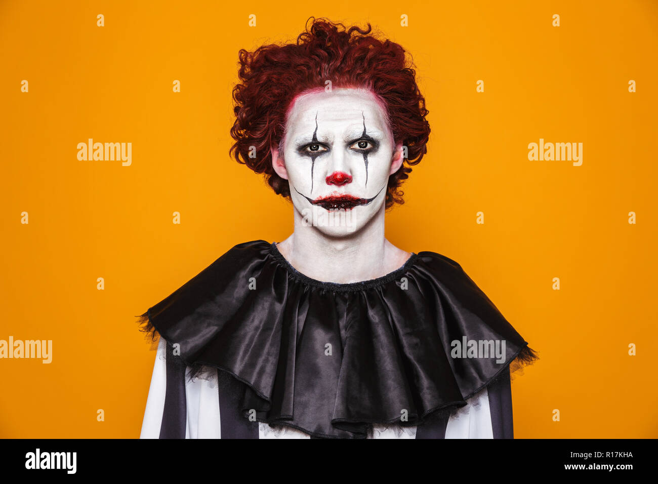 Sad clown man 20s wearing black costume and halloween makeup looking at camera isolated over yellow background Stock Photo