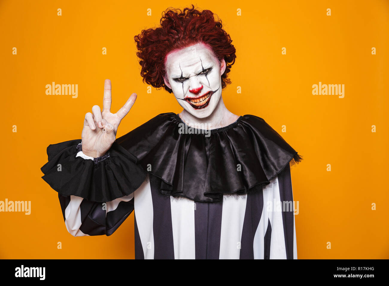 Frightening clown man 20s wearing black costume and halloween makeup looking at camera isolated over yellow background Stock Photo