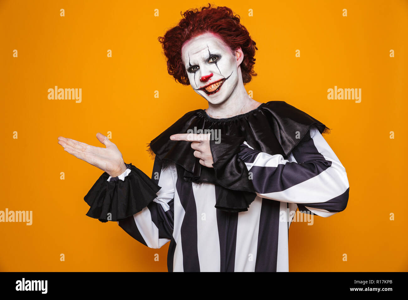 Horrific clown man 20s wearing black costume and halloween makeup holding copyspace at palm isolated over yellow background Stock Photo