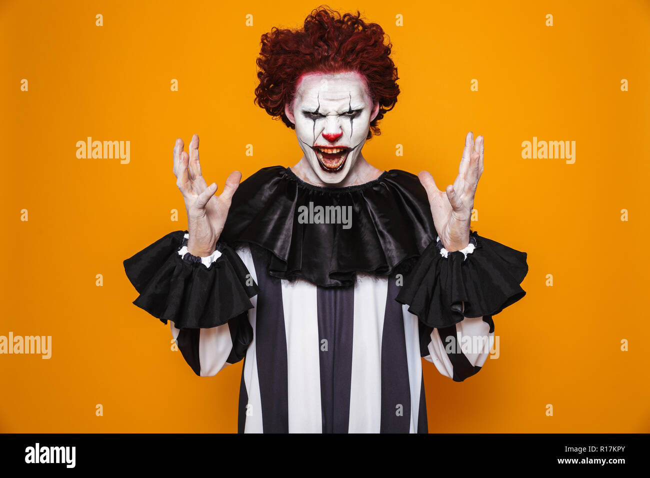 Ugly clown man 20s wearing black costume and halloween makeup looking at camera isolated over yellow background Stock Photo