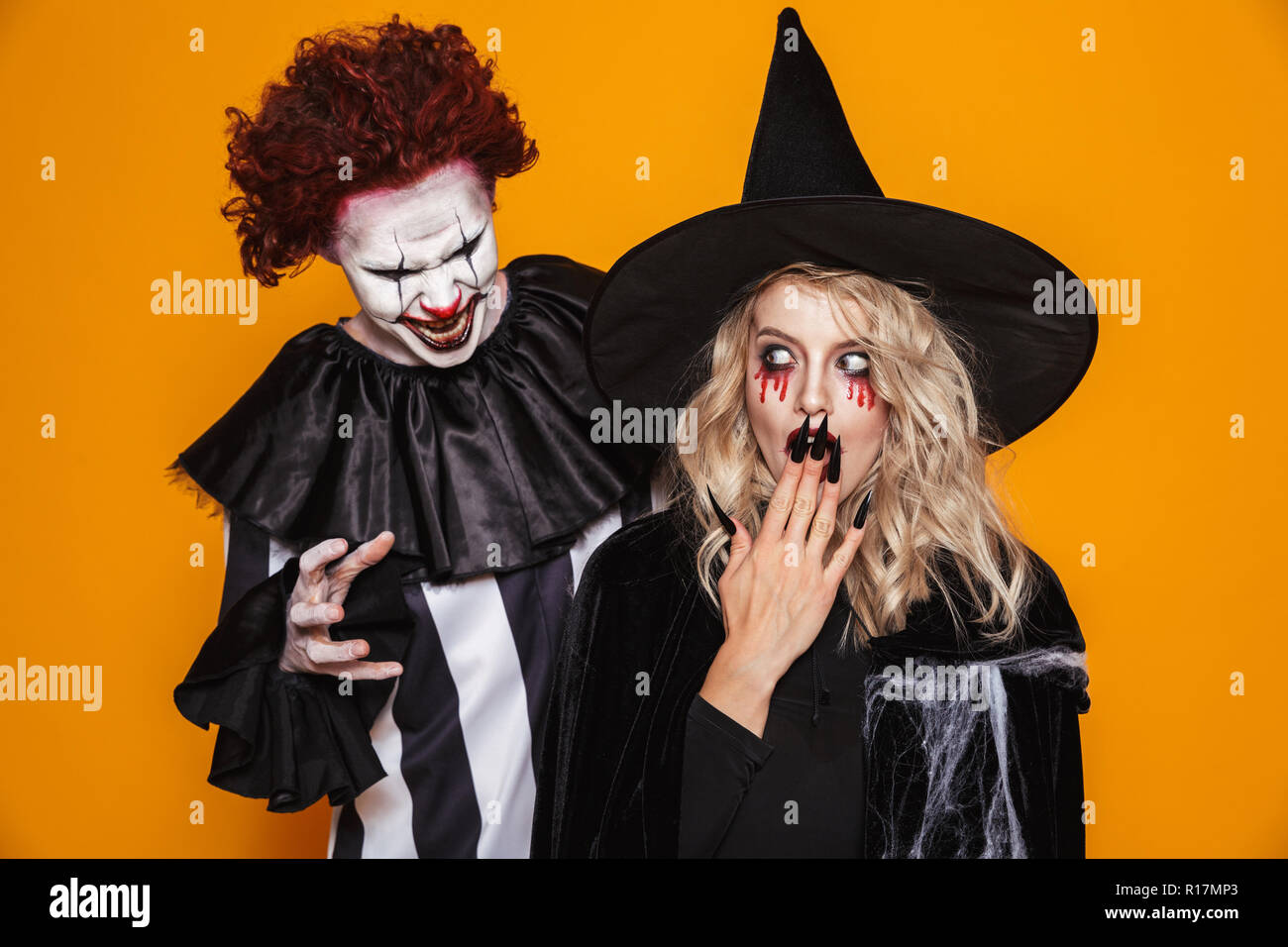 Terrifying clown with halloween make-up scary blonde witch in black hat isolated over orange Stock Photo
