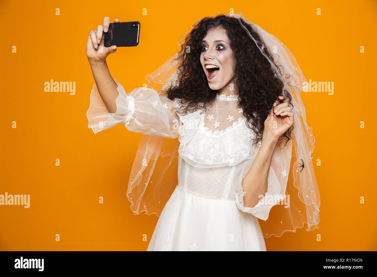 Image of horrifying bride zombie on halloween wearing wedding dress and scary makeup taking selfie on mobile phone isolated over yellow background Stock Photo