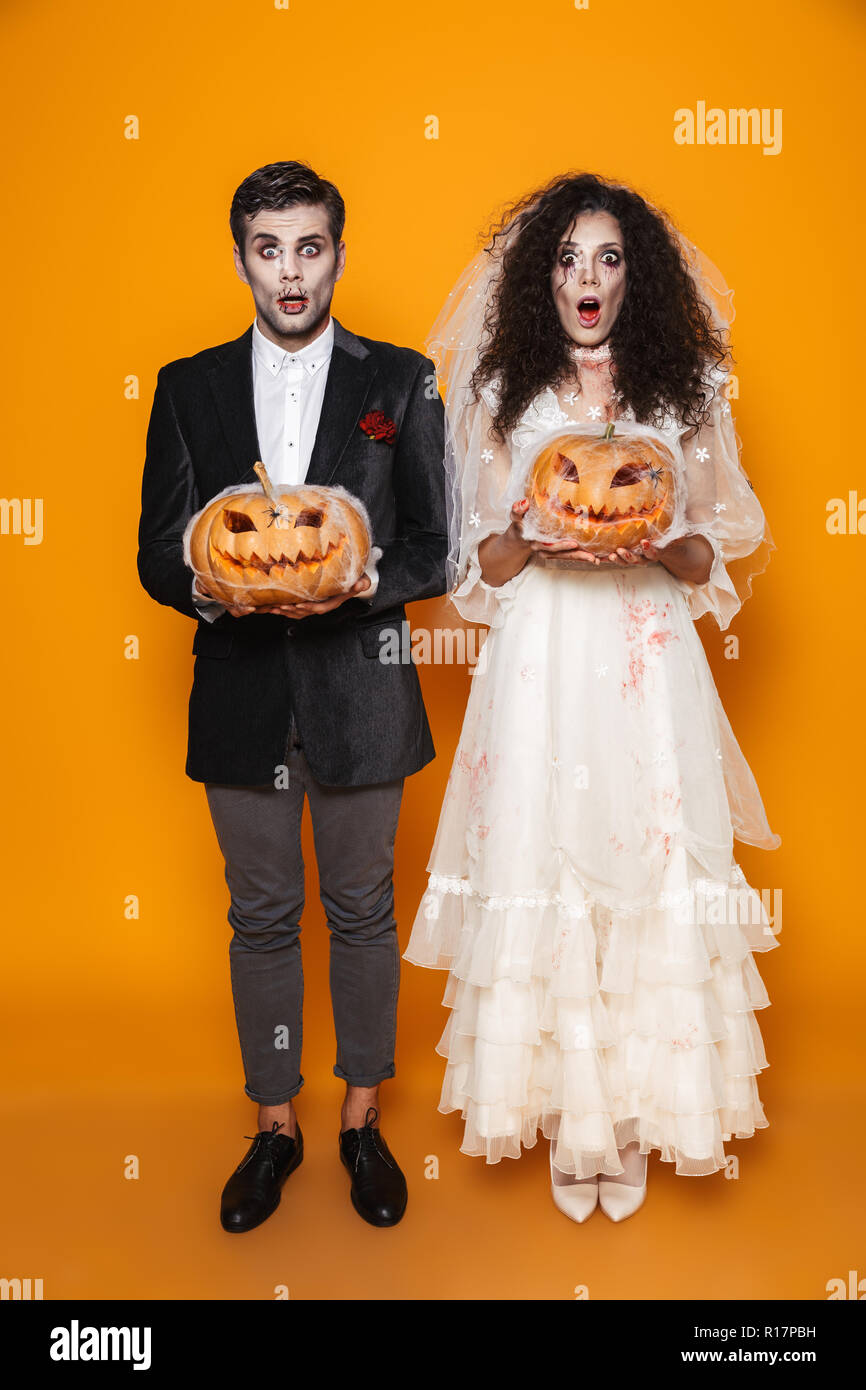 Full Length Shot Of Surprised Scary Halloween Bride And Groom In Full Length Shot Of Surprised Scary Halloween Bride And Groom In Wedding Costumes Looking Camera And Holding Pumpkin In Web Isolated Over Orange R17PBH