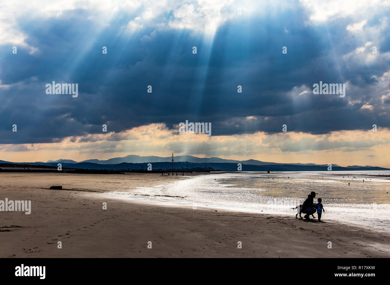 Empty beach on a gloomy moody day with sun beams coming through the stormy clouds.  Father, son & pet dog look out to sea.  Taken at Prestatyn, North  Stock Photo