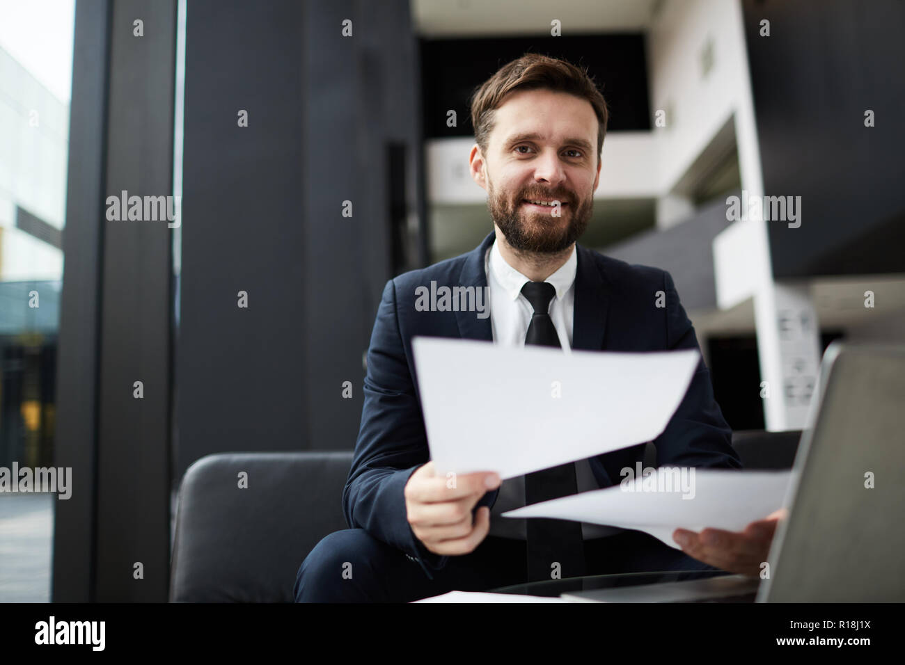 Happy bearded businessman with papers looking at you while working in modern office Stock Photo
