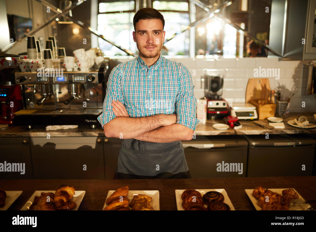 Cross-armed guy in workwear looking at you while selling fresh pastry and puff snacks in cafeteria Stock Photo