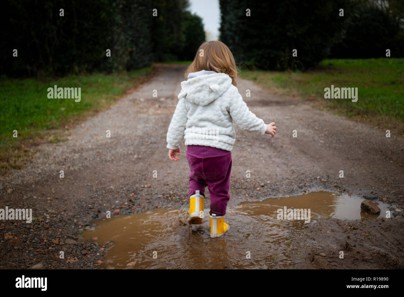 Toddler girl, back view, splashing in puddles in muddy country road with with yellow rain boots. Subject in the center. Stock Photo