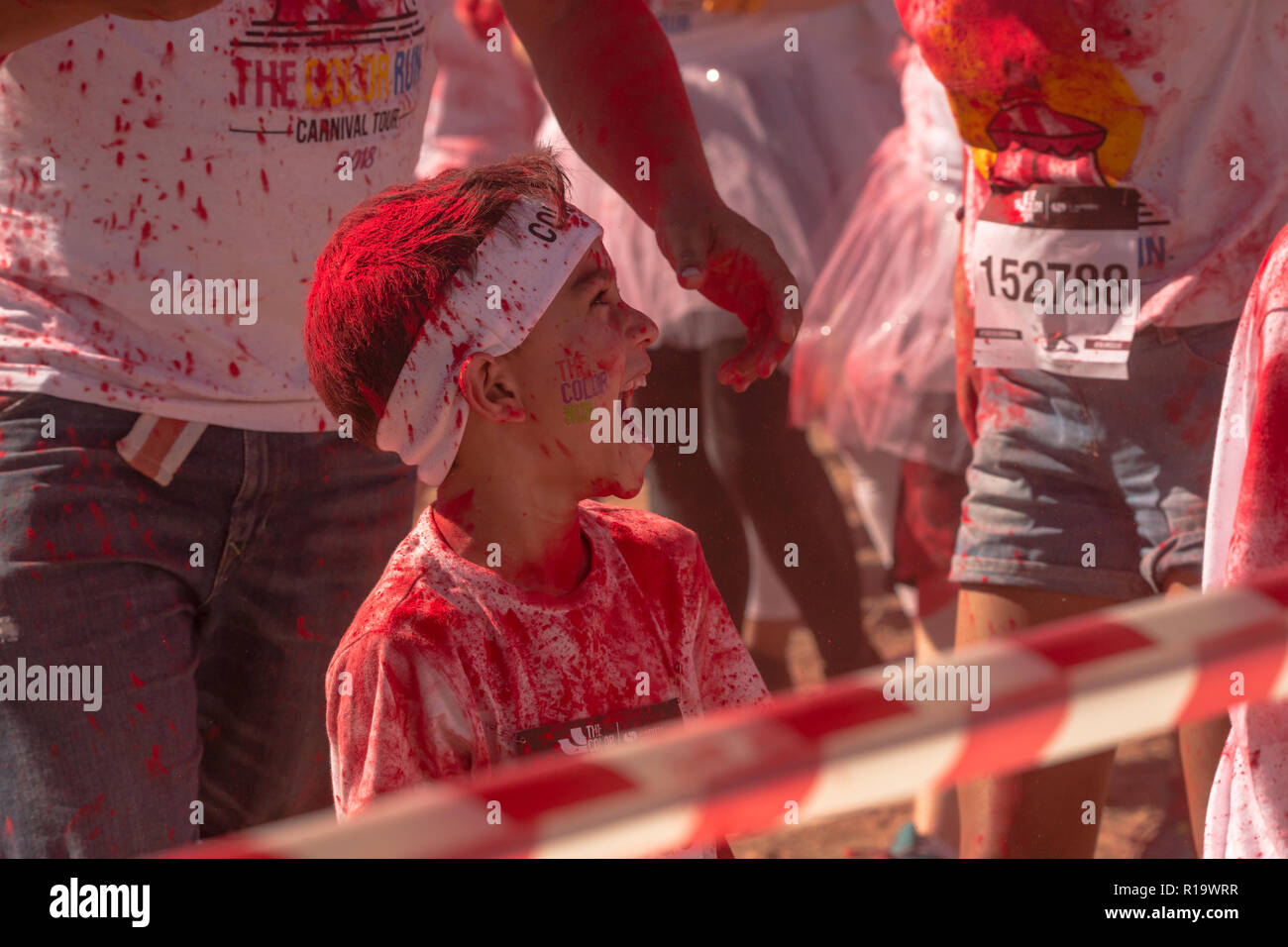 caucasian little boy about 5-7 years old laughing and full of joy covered in red powder paint and wearing a white headband surrounded by people at a Summer fun run event Stock Photo