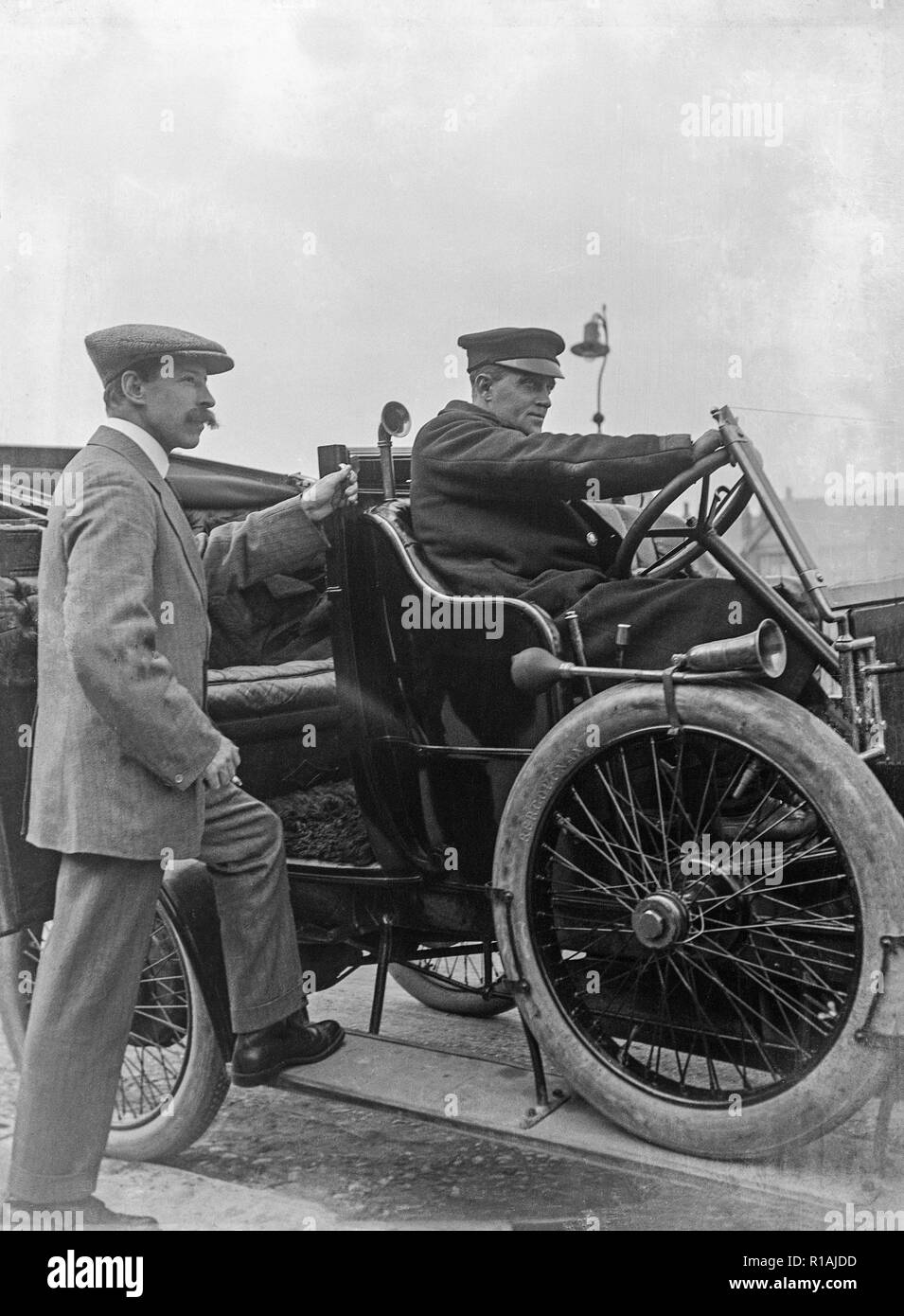 A black and white photograph taken during the early 20th century showing two men and a vintage motor car. One man behind the steering wheel, and one man standing with one foot on the running board of the car. Stock Photo
