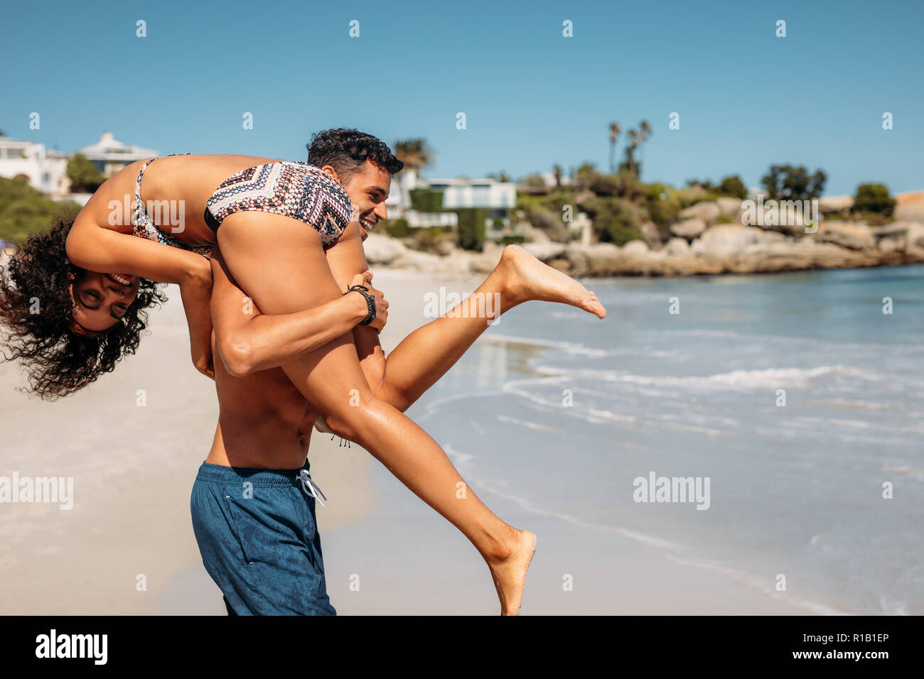 Man walking on beach carrying his girlfriend on his shoulder. Young tourist couple in romantic mood enjoying on the beach. Stock Photo
