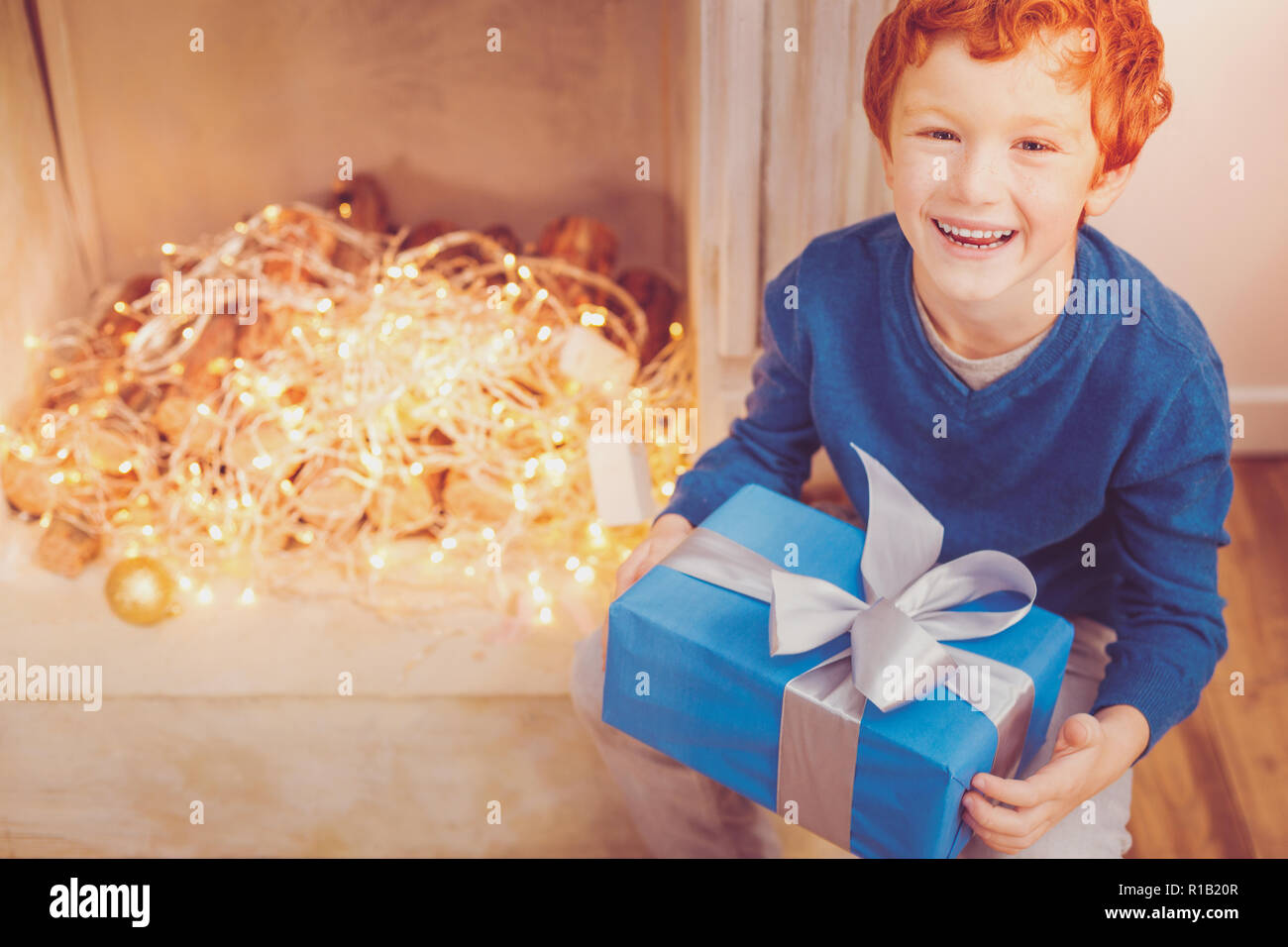 Joyful red-haired boy posing with Christmas present near chimney Stock Photo
