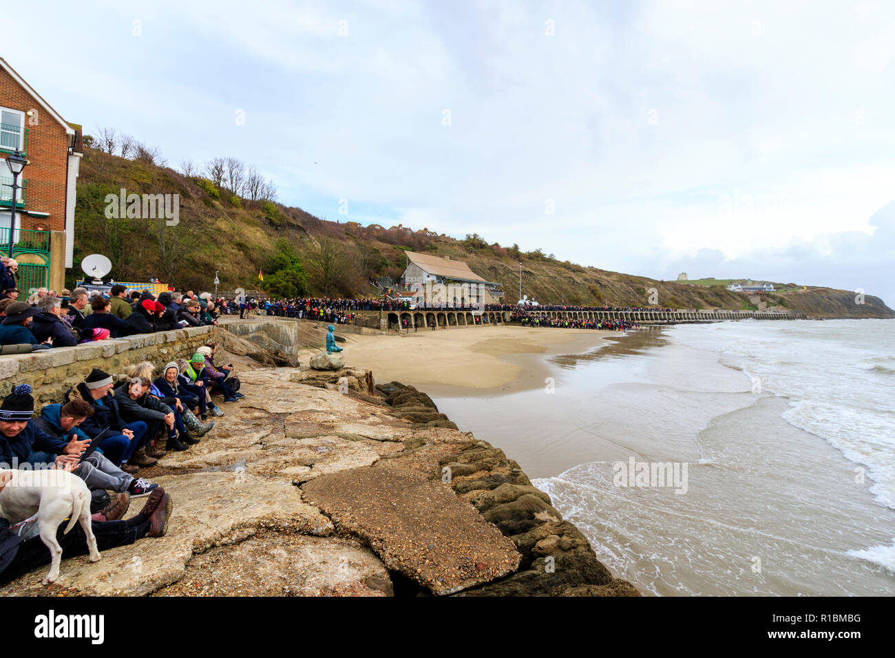 Folkestone, Kent, England. 11 November 2018. People lining the seafront, under grey skies, to watch a sand portrait of Wilfred Owen slowly been washed away by the incoming tide. 100th anniversary of the end of World war one, Remembrance Sunday event, 'Pages of The Sea'. Stock Photo