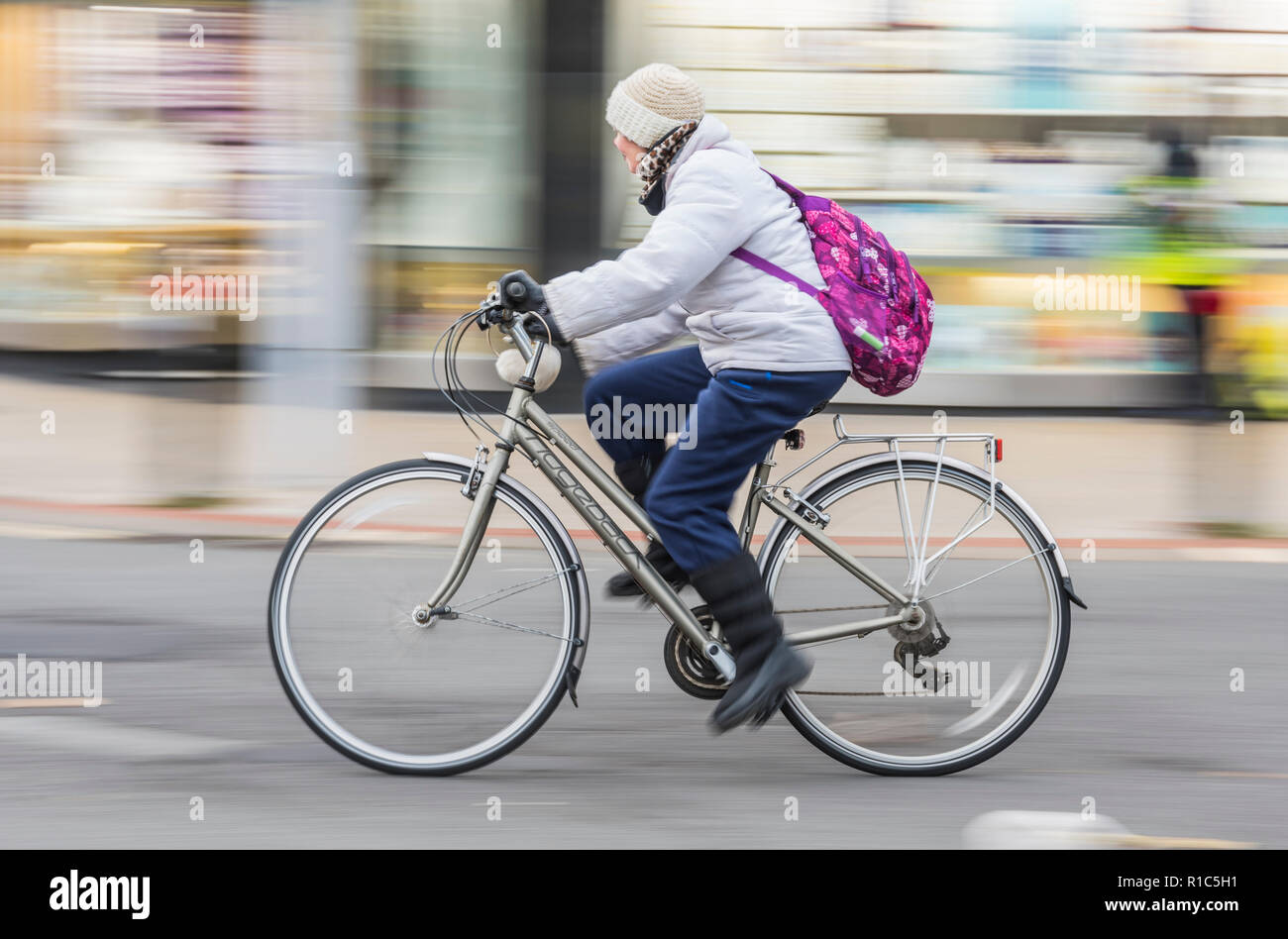 Side view of middle aged woman riding a Ridgeback bicycle wearing hat, coat & backpack, showing motion blur, UK. Female cycling. Female cyclist. Stock Photo