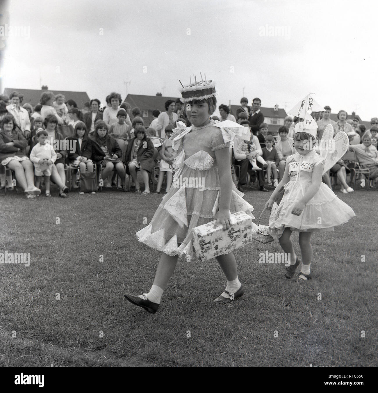 1967, historcial, outside in a fieldd, at an English village fete, two young girls dressed in costumes walk infront of the spectators in a fancy dress competition, England, UK. One of the girls has a party dress on and wearing a cake on her head, while the other is dressed as an angel and has the words Gipsy Moth IV on her, the name of the boat Sir Francis Chichester who the following month, successfuly sailed, single-handed, around the world. Her thoughts were with him. Stock Photo