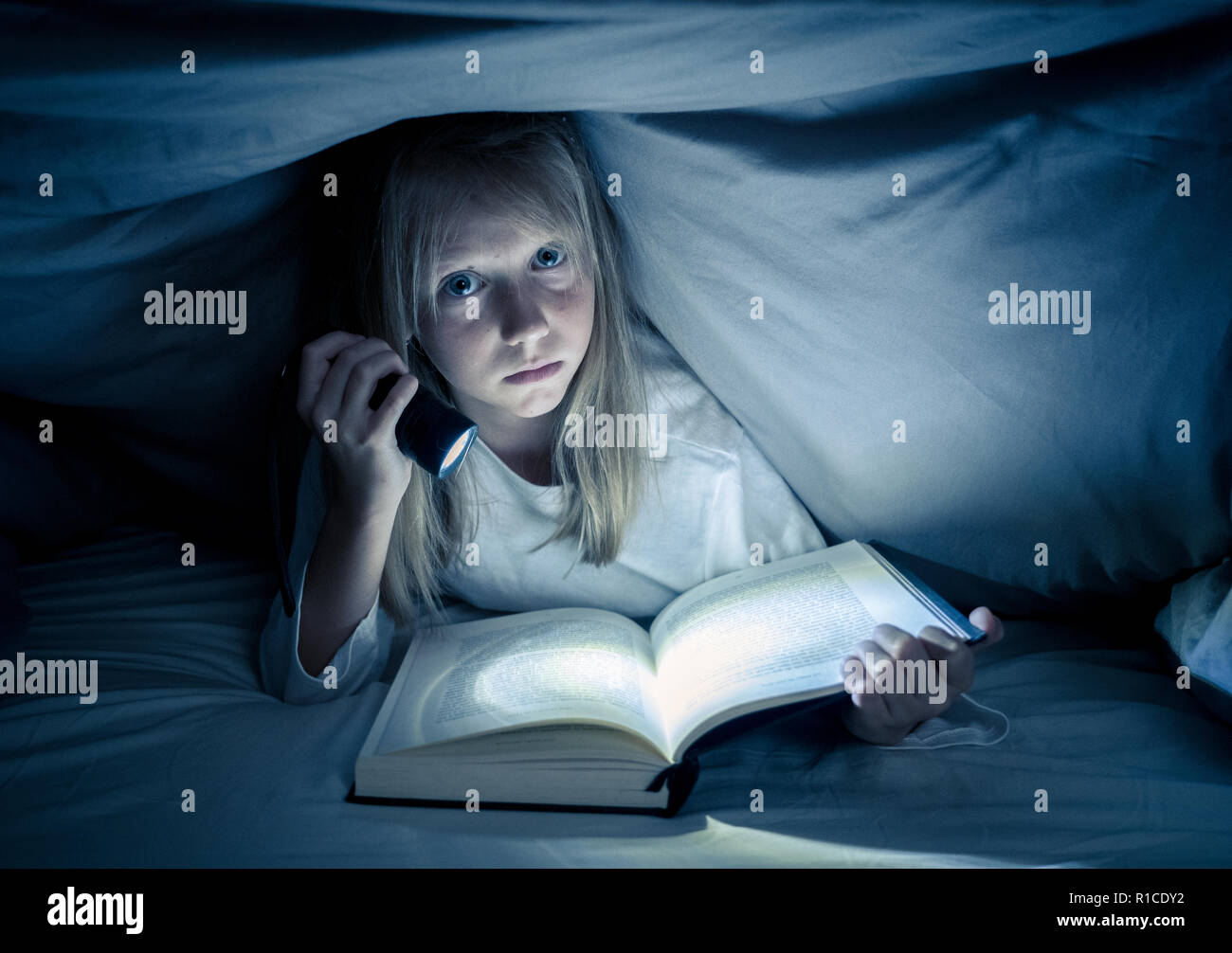 Beautiful caucasian girl lying in bed under the duvet holding a lantern reading a mystery book in the dark late at night looking frightened with a dra Stock Photo