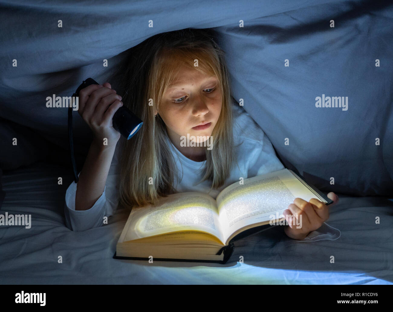 Beautiful caucasian girl lying in bed under the duvet holding a lantern reading a mystery book in the dark late at night looking frightened with a dra Stock Photo