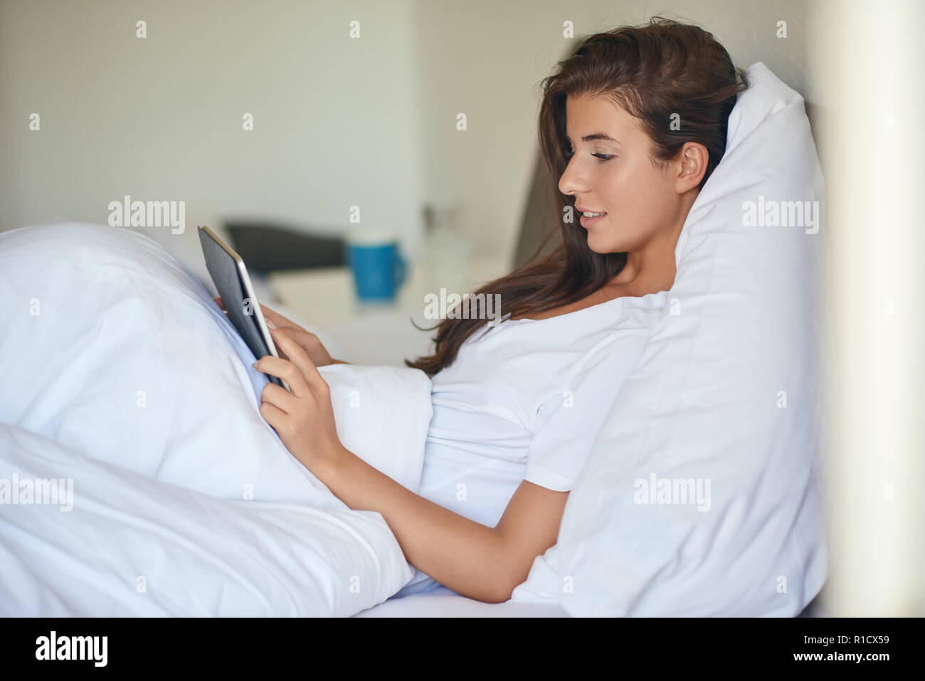 Young pretty woman with long brunette hair lying in bed under white blanket with tablet PC in her hands, looking at camera and laughing with copy spac Stock Photo