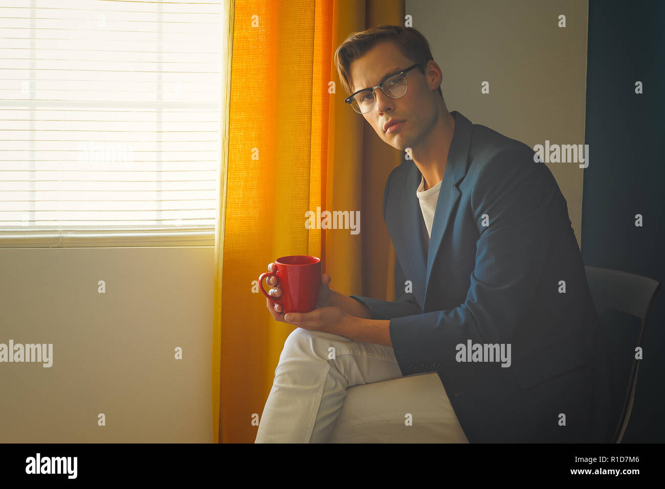 A young man, male model in blue suit and white pants,wears a pair of vintage glasses, holding a red mug. A men's vintage fashion concept. Stock Photo