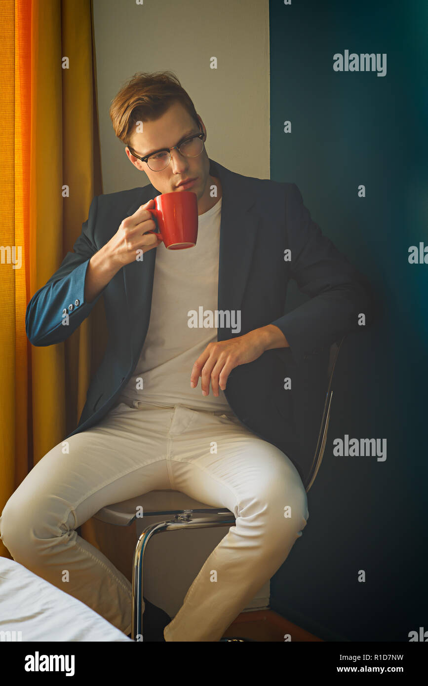 A young man, male model in blue suit and white pants,wears a pair of vintage glasses, looking away,holding a red mug. A men's vintage fashion concept. Stock Photo