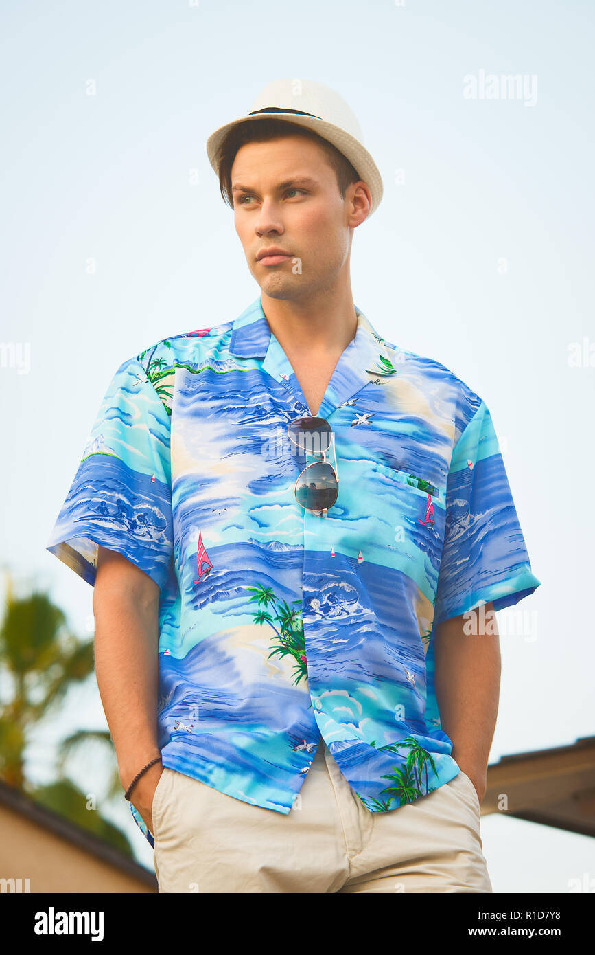 A handsome male model posing in Summer shirt outdoor against a blue sky. He wears a white Fedora hat. Stock Photo