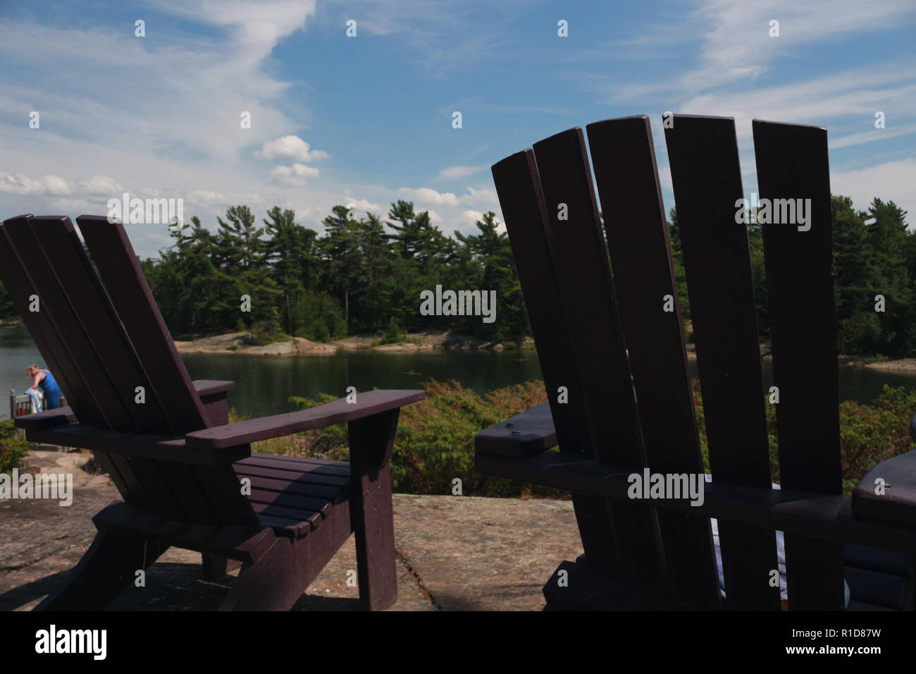 Two Muskoka chairs sitting on a wood dock facing a lake. Across the ...