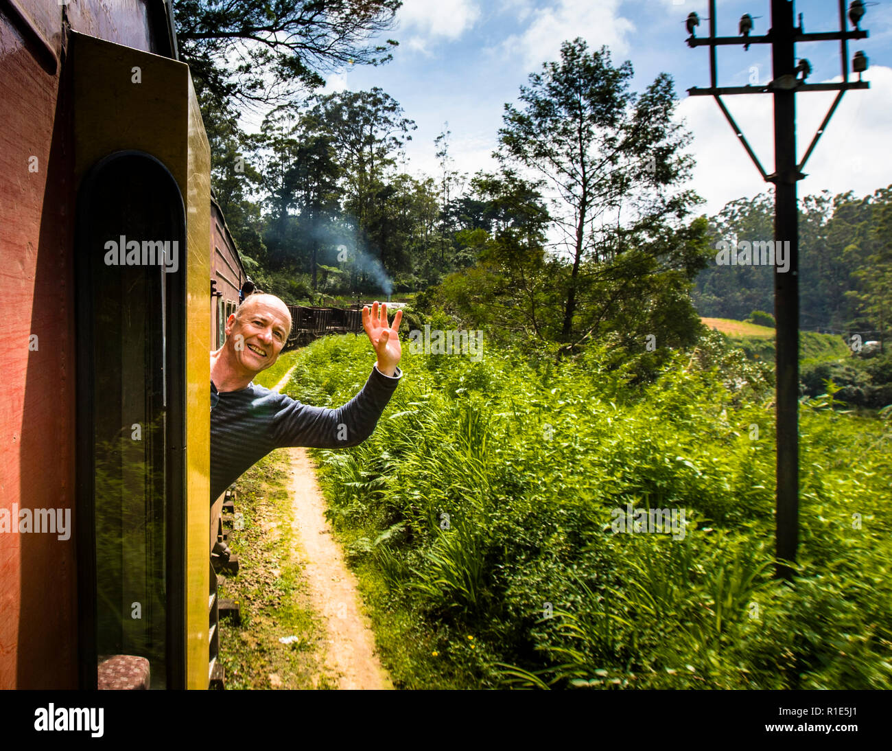 Photographer Georg Berg leans out of the open door of a train carriage, laughing and waving, near Ambewela railway station between Nanu Oya and Ella, Sri Lanka, as the steam of an approaching locomotive rises in the background. During the ride, Georg Berg handed his camera to a fellow passenger so he could be photographed himself. Stock Photo