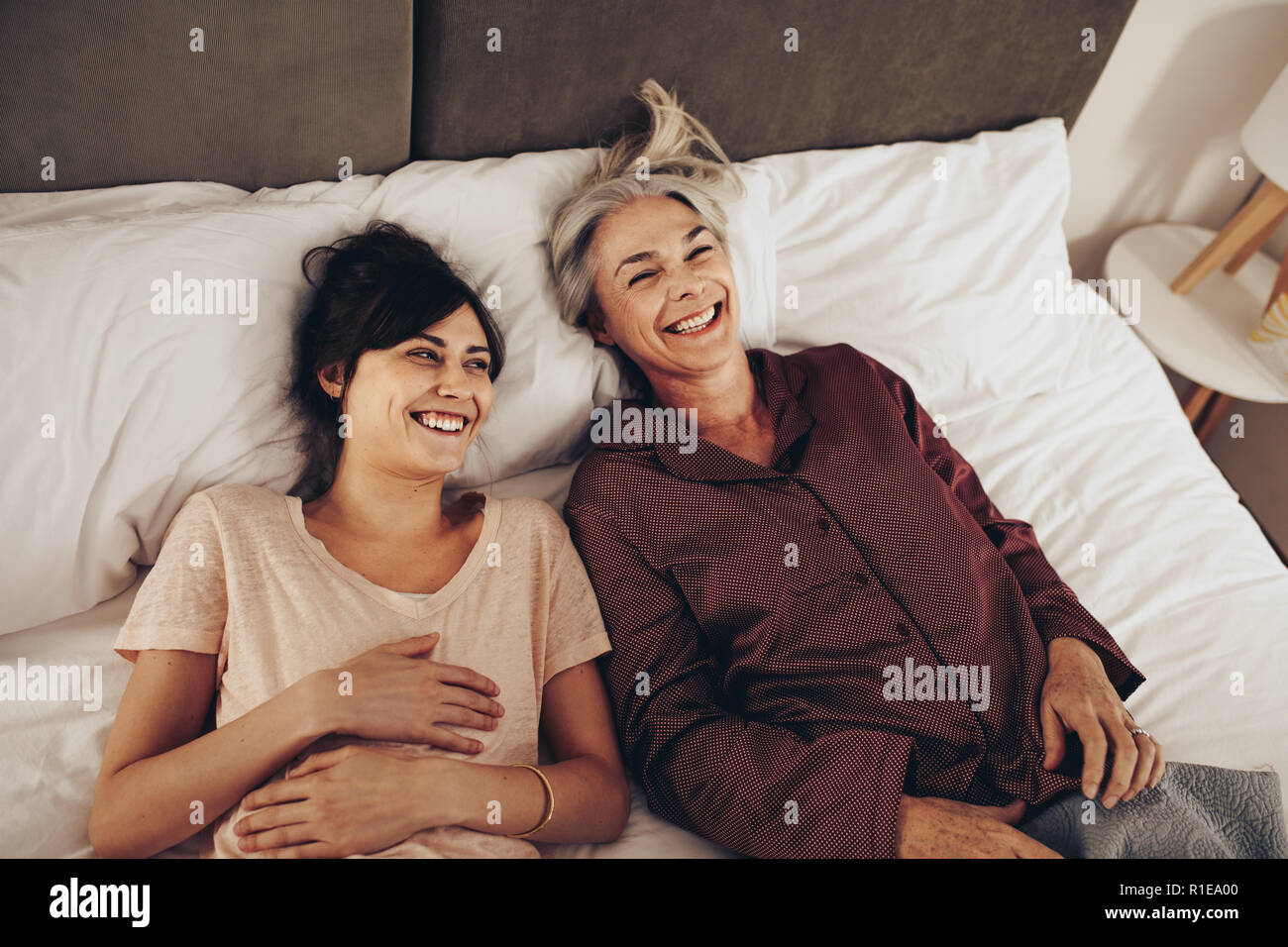 Mother and daughter lying on bed and laughing. Woman spending time talking with her elderly mother lying on bed at home. Stock Photo