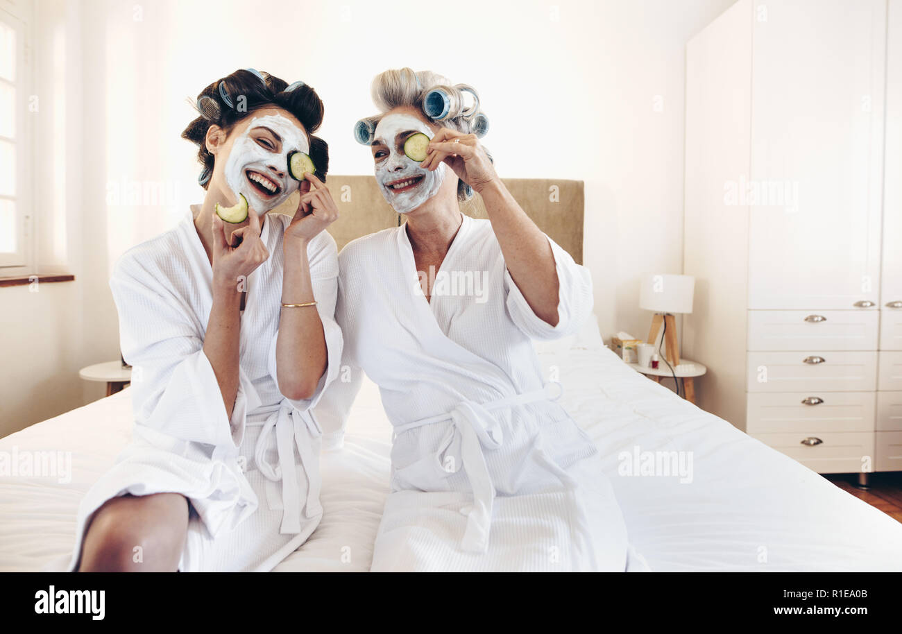 Smiling mother and daughter in bathrobes with face pack holding slices of cucumber to eyes. Happy women sitting on bed with rollers pinned on hair. Stock Photo