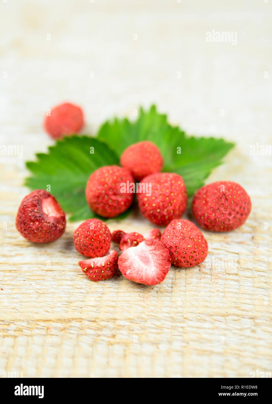 Freeze dried whole and pieces of strawberries on green strawberry leaf, healthy snack full of vitamins and nutrition on natural wooden background. Stock Photo