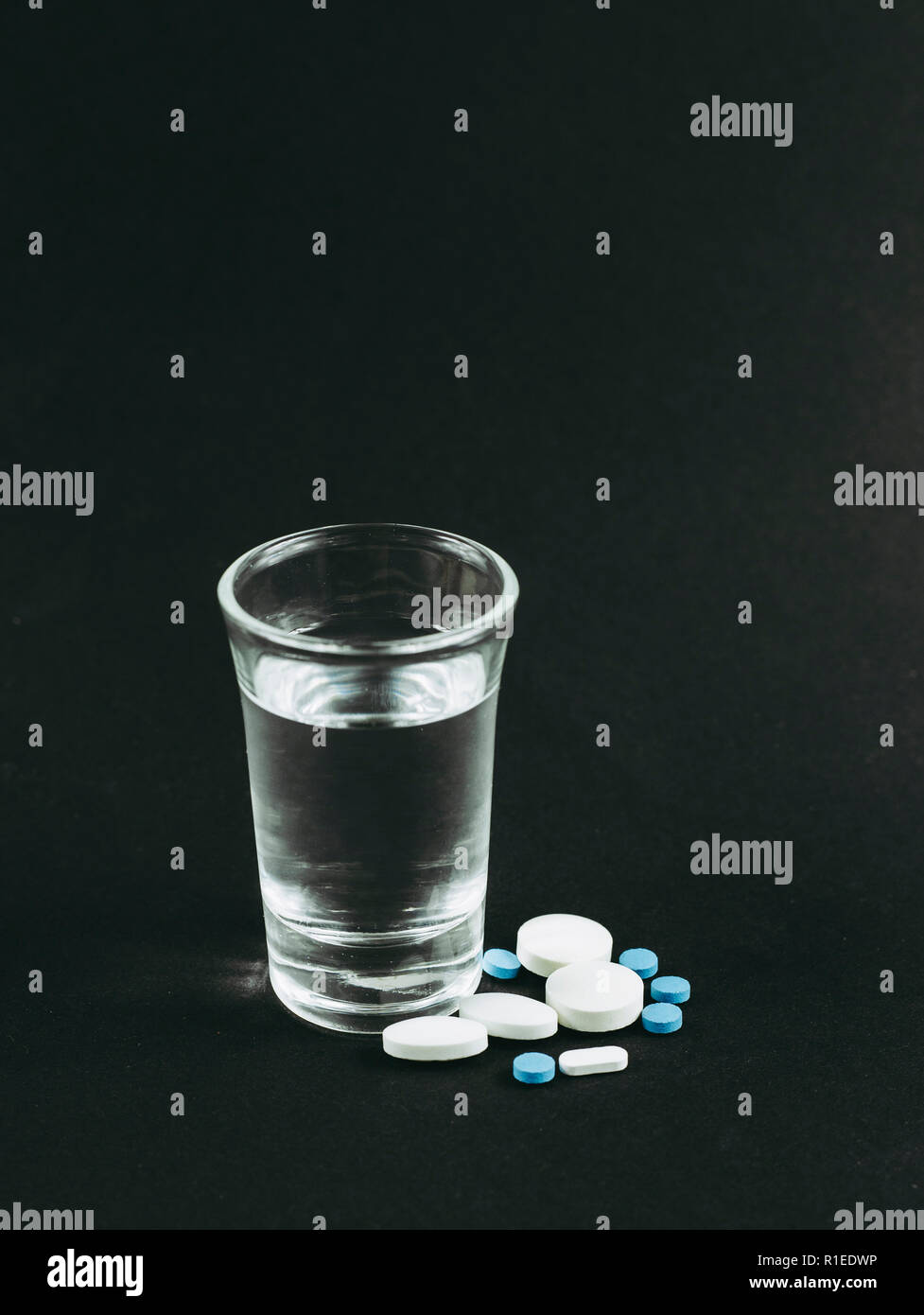 Mixing alcohol with drugs pills is bad dangerous concept. Glass of vodka with pills tablets drugs on black background. Stock Photo