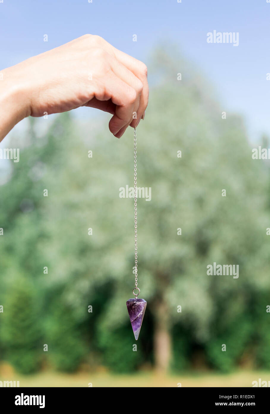 Purple natural Amethyst crystal quartz pendulum hanging on chain on green outdoors nature background. Stock Photo