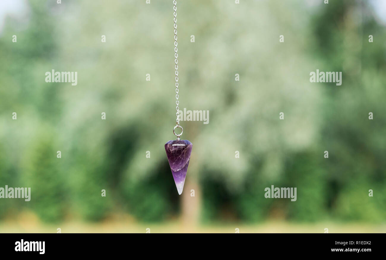 Purple natural Amethyst crystal quartz pendulum hanging on chain on green outdoors nature background. Stock Photo