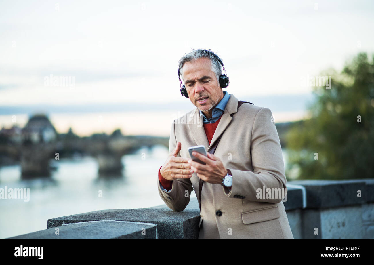 Mature businessman with headphones and smartphone standing by river, singing. Stock Photo