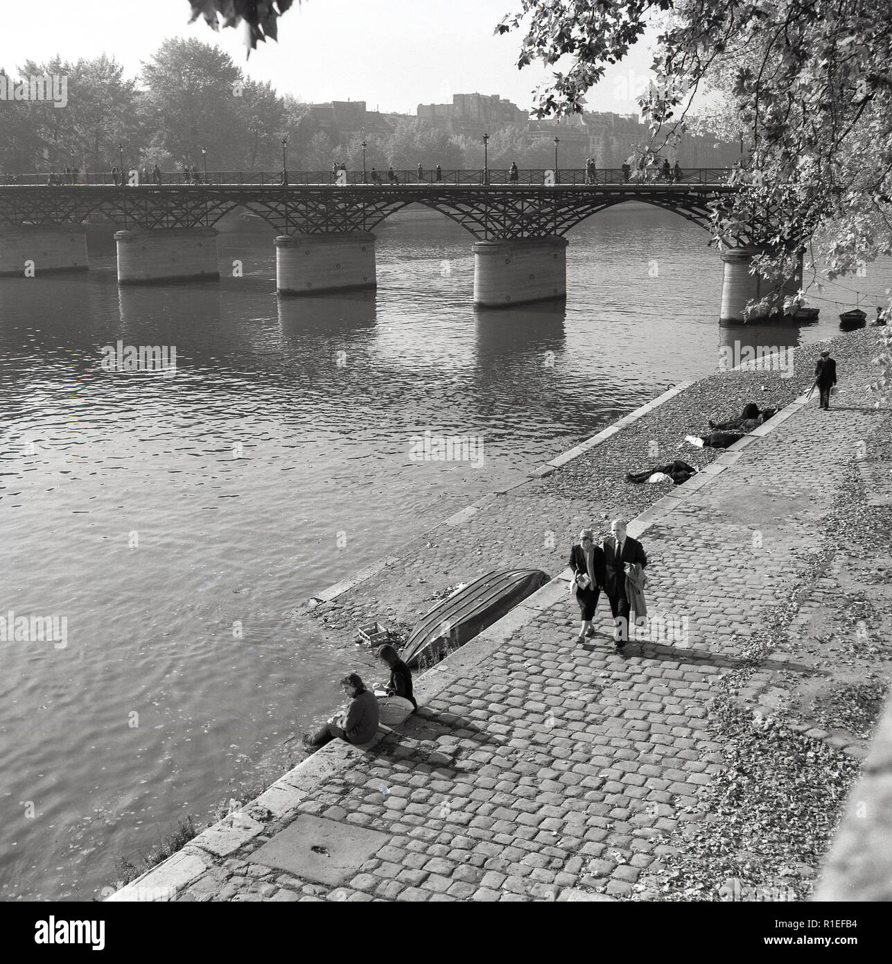 1950s, historical, daytime and a Parisian couple walk along a cobblestone path beside the river Seine, Paris, France. Stock Photo